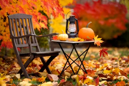 A photo of a cozy outdoor setting with a wooden chair and a small table. On the table, there is a lantern, a pumpkin, and a few autumn leaves. The background is a garden with vibrant autumn leaves in shades of orange, yellow, and red. The ground is covered with fallen leaves. The overall image has a warm color scheme palette.